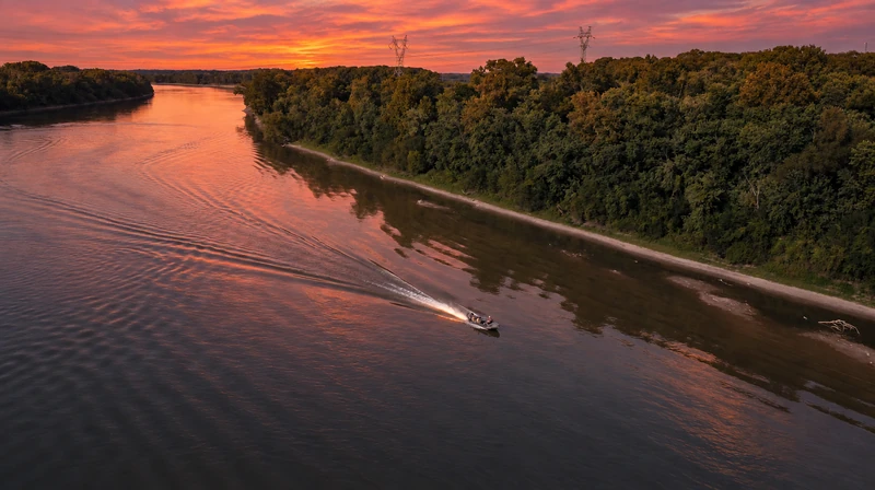 A motorboat and its wake on a wooded river beneath an orange sunset sky